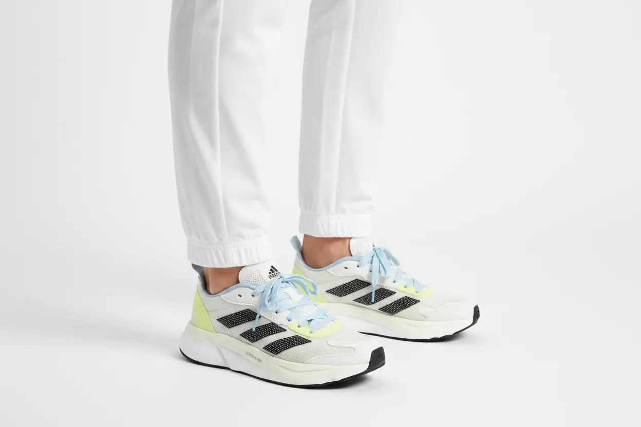 White Adidas sneakers with black three-stripes, light blue laces, and neon yellow-green accents, shown on a person wearing white pants against a light gray studio background.