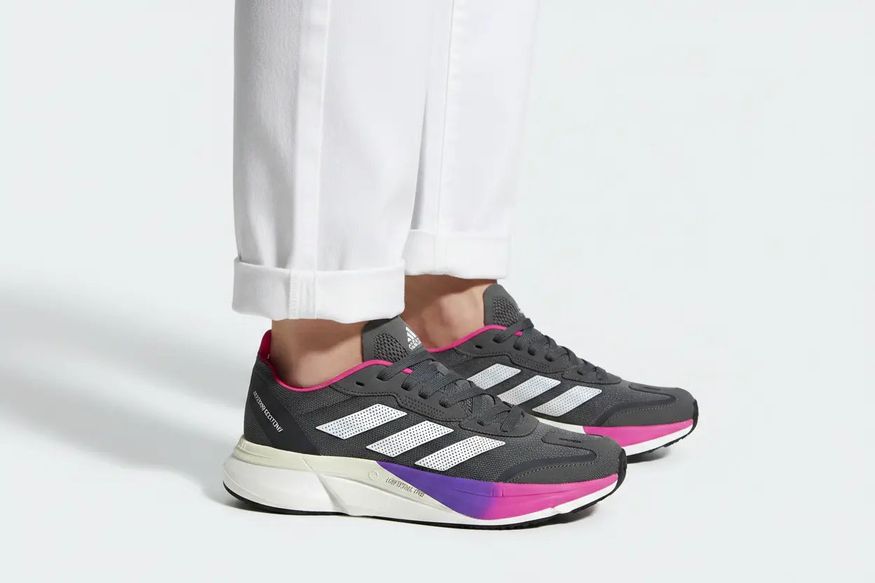 A pair of Adidas running shoes in dark gray with white three-stripes, a white midsole, and a vibrant pink and purple gradient outsole, shown on a model wearing white pants against a light gray background.
