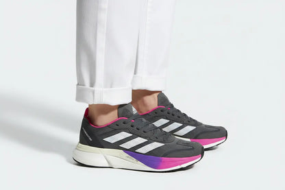 A pair of Adidas running shoes in dark gray with white three-stripes, a white midsole, and a vibrant pink and purple gradient outsole, shown on a model wearing white pants against a light gray background.