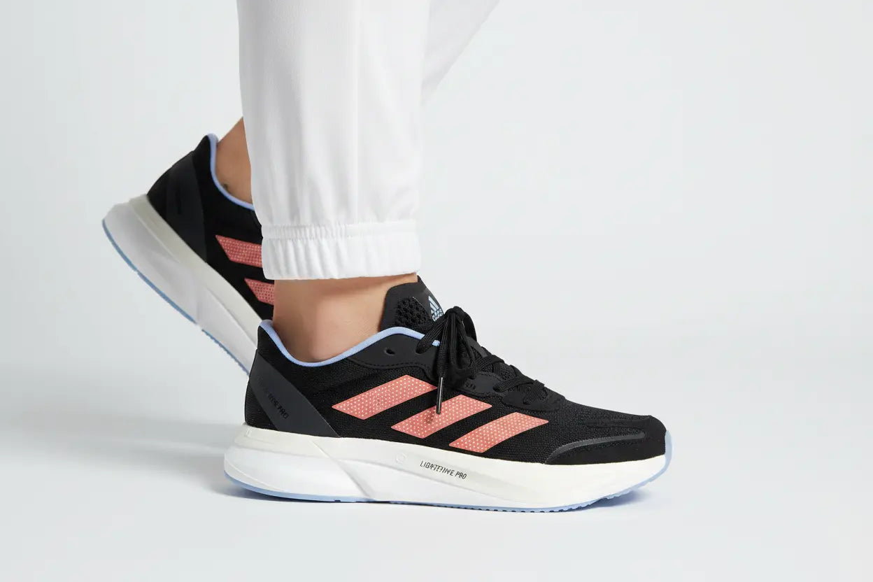 A pair of Adidas running shoes in black and coral pink, shown on a model wearing white pants against a light gray studio background.