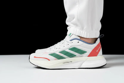 A white Adidas Lightstrike Pro sneaker with green three-stripes, a coral-red heel accent, and blue lining, shown on a person's foot wearing white pants against a black and white studio background.