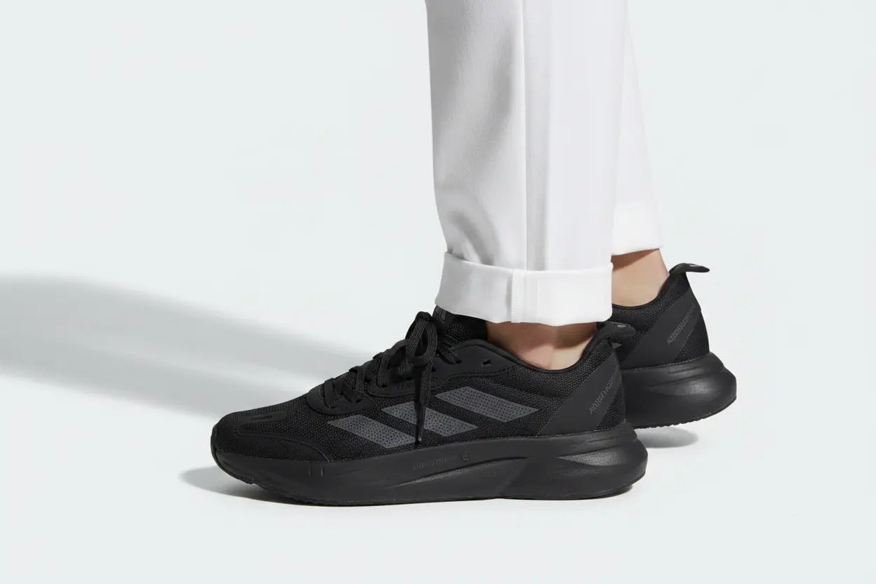 Black Adidas sneakers with dark gray three-stripes, shown on a model wearing white cuffed pants against a light gray studio background.